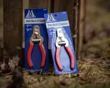 Två förpackningar med Millers klotång, med röda handtag och metallskär, står lutade mot ett trästaket.