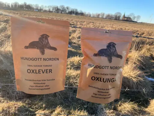 Två påsar med hundgodis, 100% svensk torkad nötlunga och oxlever, hänger på ett taggtrådsstängsel i ett soligt, torrt landskap.