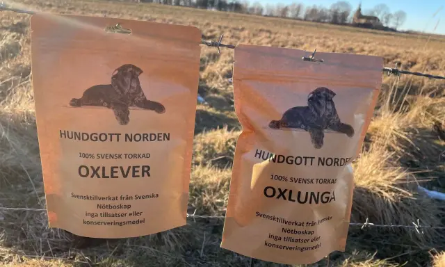 Två påsar med hundgodis, 100% svensk torkad nötlunga och oxlever, hänger på ett taggtrådsstängsel i ett soligt, torrt landskap.