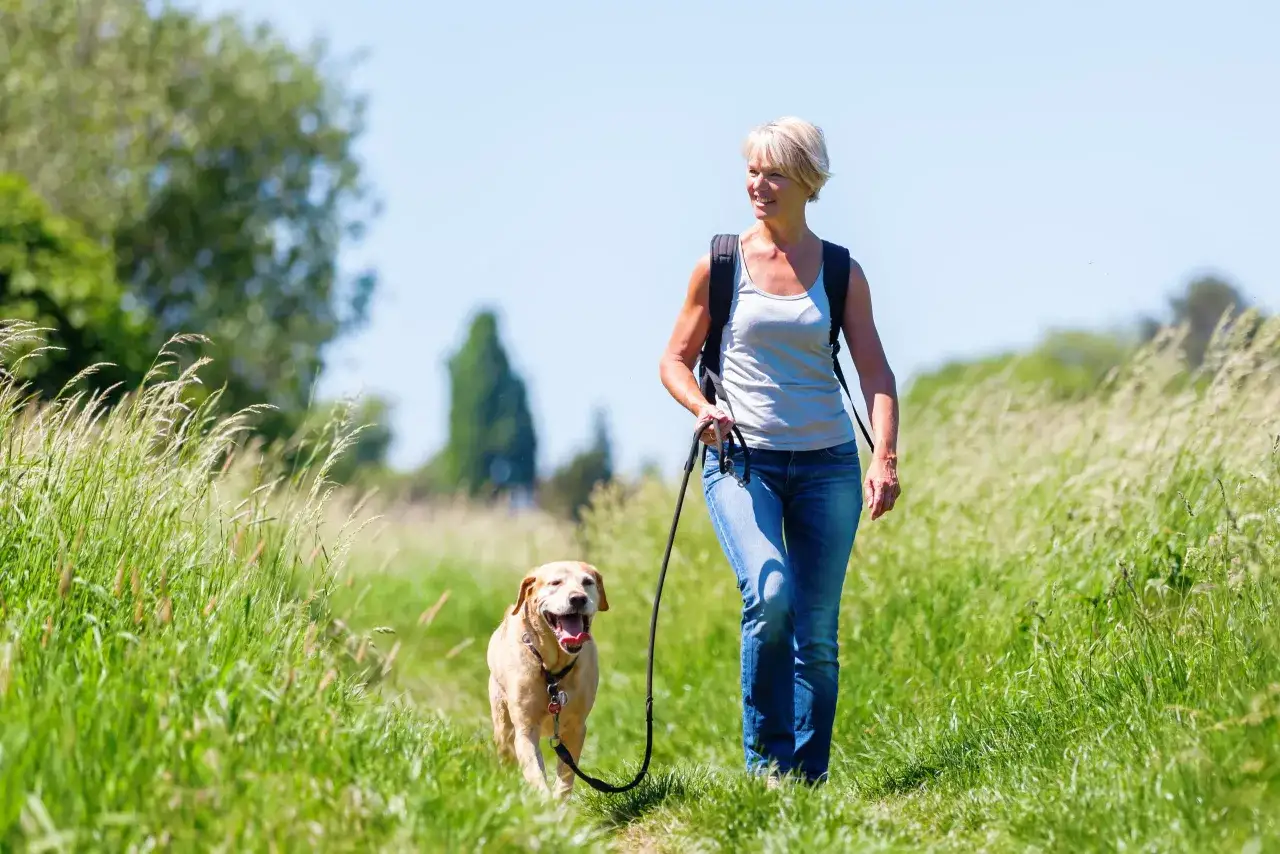 Kvinna med ryggs&auml;ck och hund p&aring; promenad i gr&auml;set. Hunden har ett halsband med koppel.